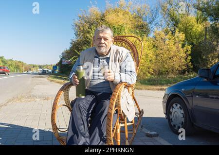 Portrait of ukrainian senior man sitting in rrocking chair on roadside and holding vacuum bottle getting ready to drink tea at sunny autumnal day Stock Photo