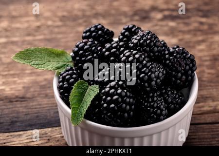 Blackberries in a white bowl with mint leaves on a wooden background close-up. Stock Photo