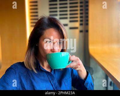 Caucasian woman sipping coffee from green cup indoors in cafe. Close up. Beautiful middle-aged lady drinking coffee resting in coffee shop and looking Stock Photo