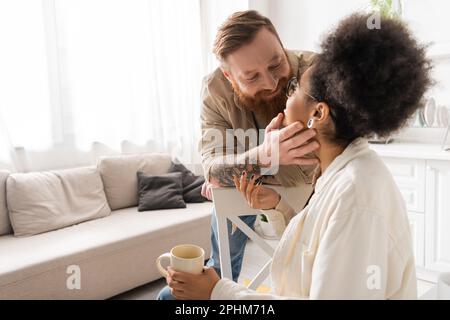 Smiling tattooed man touching face of african american girlfriend with coffee cup in kitchen,stock image Stock Photo