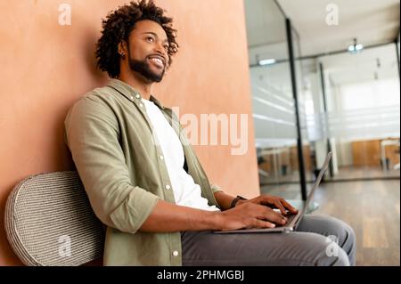 Smiling optimistic young male entrepreneur in casual wear, sitting on the chair, typing on keyboard, freelancer professional is feeling ready to work and start new projects Stock Photo