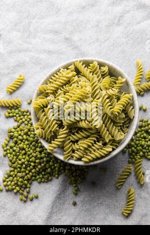 Mung bean fusilli pasta on a gray textile background. Bowl with raw pasta and green mung bean. Gluten free pasta. Stock Photo