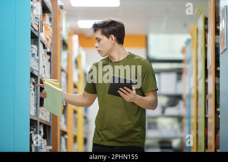 University student with digital tablet taking books from shelves in library Stock Photo