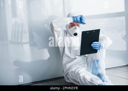 Tired doctor scientist in lab coat, defensive eyewear and mask sits and takes break Stock Photo
