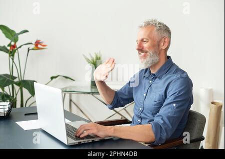 Cheerful mature grey-haired man using laptop for video call sitting at the desk in the office, senior male looks at the webcam, smiling and waving. Virtual meeting concept. Side view Stock Photo