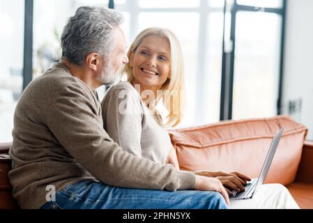 Cheerful and happy middle-aged spouses spending time with a laptop at home. Handsome grey-haired man and beautiful good-looking woman sit on the sofa, using laptop and smile. Side view Stock Photo
