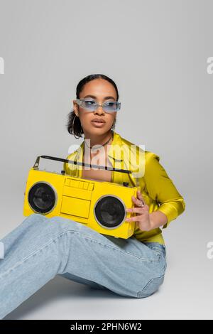 stylish african american woman in blue sunglasses sitting and holding retro boombox on grey,stock image Stock Photo