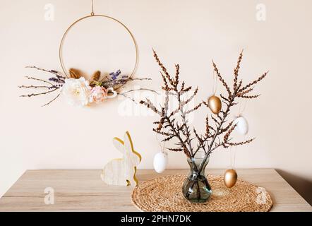 Home Easter decor. Handmade spring round flower garland hanging on wall, tree branches on vase with eggs hanging from it. Nice warm beige earthly tone Stock Photo