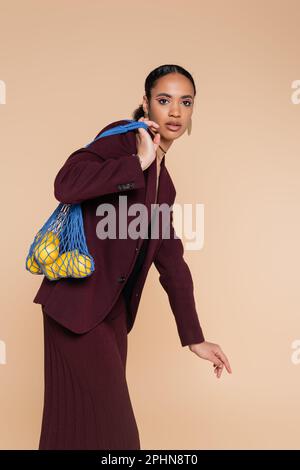 stylish african american woman in burgundy blazer holding net bag with fresh lemons isolated on beige,stock image Stock Photo