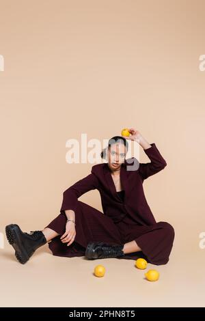 young african american woman in stylish burgundy suit sitting around fresh lemons on beige,stock image Stock Photo