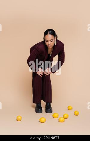 stylish african american woman in burgundy suit standing and looking at fresh lemons on beige,stock image Stock Photo
