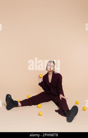 stylish african american woman in burgundy suit sitting around fresh lemons on beige,stock image Stock Photo