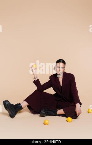 stylish african american model in maroon suit sitting around fresh lemons on beige,stock image Stock Photo