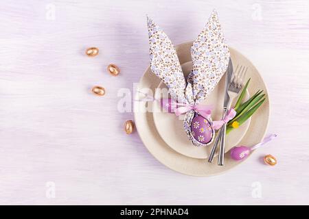 Easter table setting. White plate with a napkin folded in the shape of a rabbit, Easter and chocolate eggs on a pink background. Happy Easter holiday Stock Photo