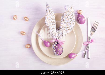 Easter table setting. White plate with a napkin folded in the shape of a rabbit, Easter and chocolate eggs on a pink background. Happy Easter holiday Stock Photo