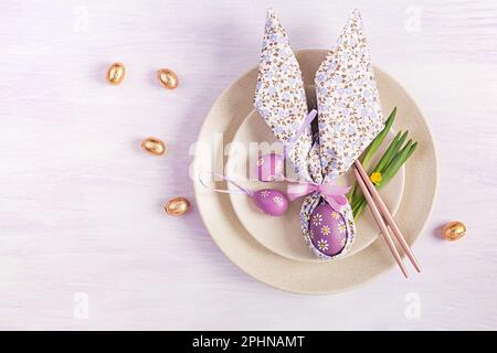 Easter table setting. White plate with a napkin folded in the shape of a rabbit, Easter and chocolate eggs on a pink background. Happy Easter holiday Stock Photo