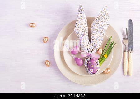 Easter table setting. White plate with a napkin folded in the shape of a rabbit, Easter and chocolate eggs on a pink background. Happy Easter holiday Stock Photo