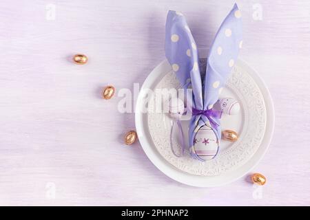 Easter table setting. White plate with a napkin folded in the shape of a rabbit, Easter and chocolate eggs on a pink background. Happy Easter holiday Stock Photo