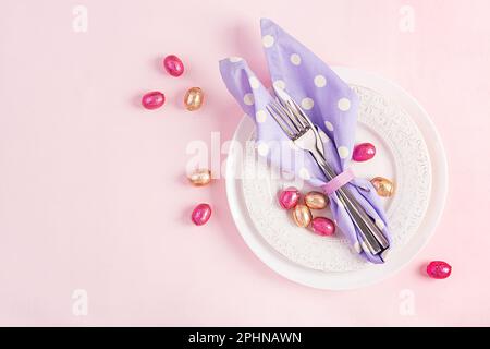Easter table setting. White plate with a napkin folded in the shape of a rabbit, Easter and chocolate eggs on a pink background. Happy Easter holiday Stock Photo