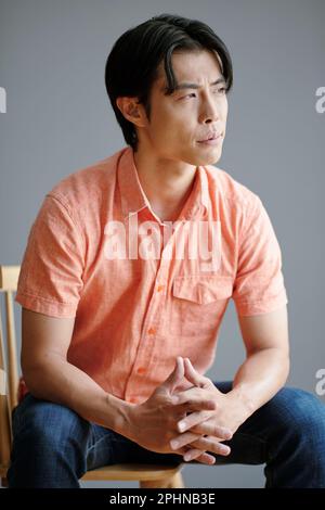 Pensive young Asian man sitting on chair and looking away Stock Photo