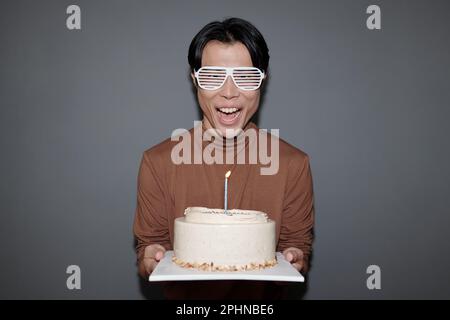 Excited young man in party glasses holding birthday cake with one candle Stock Photo