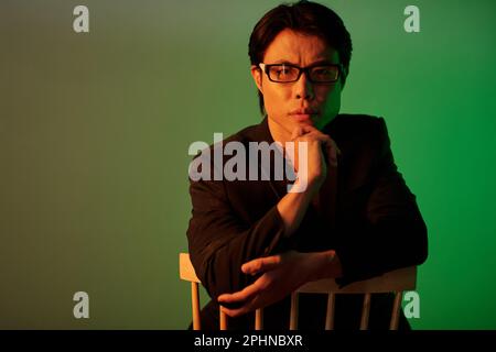 Portrait of frowning serious Asian man in glasses sitting on chair and looking at camera Stock Photo
