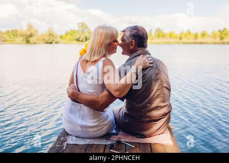 Senior family couple relaxing by summer lake. Man and woman enjoying natural landscape and hugging sitting on pier Stock Photo