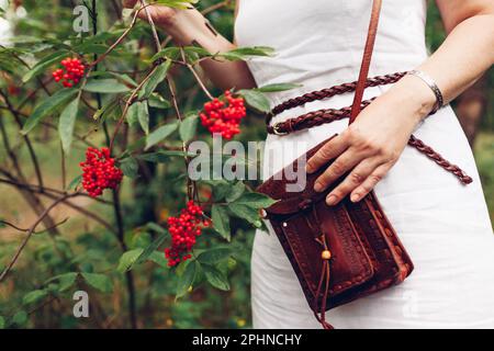 Close up of brown handmade leather purse. Stylish woman wearing white dress, belt, holding handbag by red berries outdoors. Casual boho female accesso Stock Photo