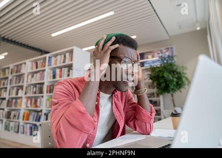 Unhappy African American student guy sitting in library looking at laptop receiving bad test results Stock Photo