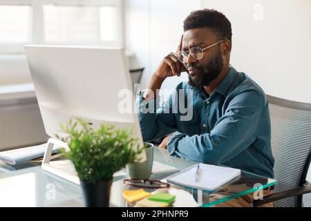 Thoughtful black male ceo working on computer in office, sitting at workplace and thinking about business project Stock Photo