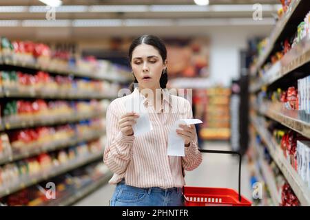 Portrait of young amazement Caucasian woman by high prices in supermarket. Shocked millennial holds receipts of purchase. In background, rows of shelv Stock Photo