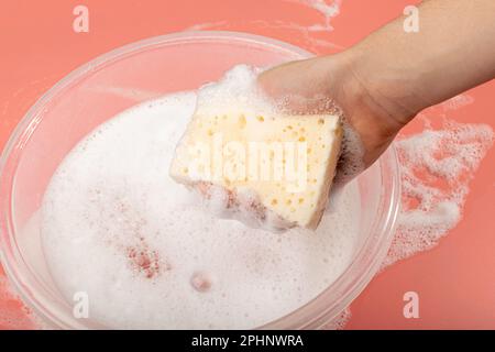 A Basin with Soapy Water, Hand Holds Soapy Sponge with Foam on Pink Background, Copy Space Stock Photo