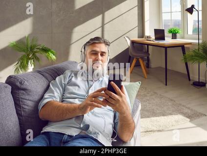 Bearded man in headphones listening to music, lying on the sofa having rest at home. Stock Photo