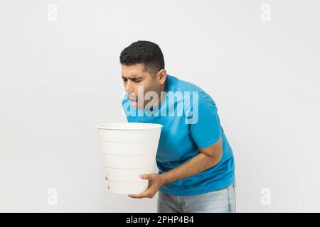 Portrait of sick unhealthy ill unshaven man wearing blue T- shirt standing suffering stomachache, feels nausea and vomits, holding bin in hands. Indoor studio shot isolated on gray background. Stock Photo