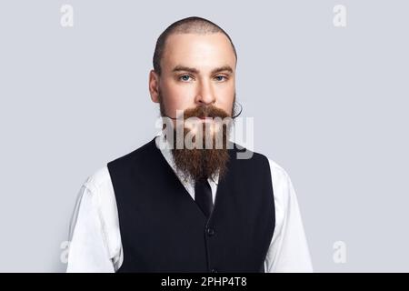 Portrait of serious attentive man with beard and mustache standing looking at camera with strict facial expression, being in bad mood. Indoor studio shot isolated on gray background. Stock Photo