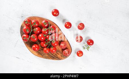 Vibrant small red tomatoes with green vines on wooden chopping board, white stone table under, view from above Stock Photo