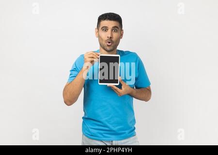 Portrait of astonished shocked unshaven man wearing blue T- shirt standing showing table t with black empty display, copy space for advertisement. Indoor studio shot isolated on gray background. Stock Photo
