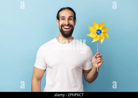 Portrait of cheerful childish man with beard wearing white T-shirt holding paper windmill, pinwheel toy on stick, playing with paper toy. Indoor studio shot isolated on blue background. Stock Photo