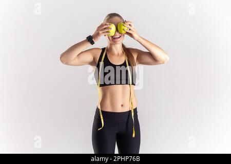 Portrait of funny athletic blonde woman standing with green apples in hands, covering eyes with fresh fruit, wearing black fitness clothing. Indoor studio shot isolated on gray background. Stock Photo