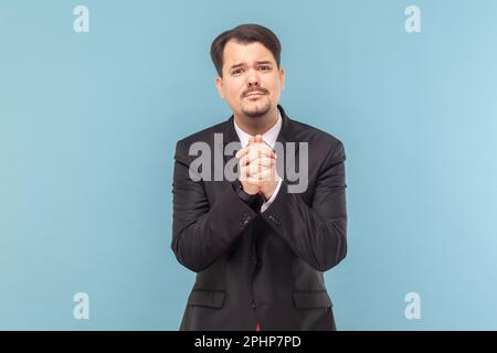 Portrait of sad upset worried man with mustache standing and pleading, looking at camera, asking to forgive him, wearing black suit with red tie. Indoor studio shot isolated on light blue background. Stock Photo