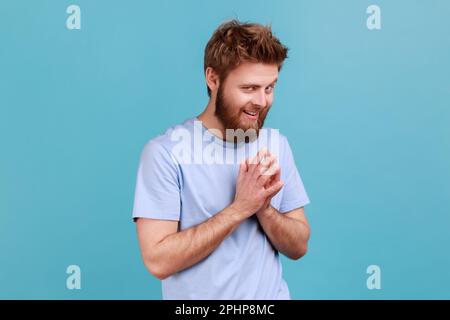 Portrait of cunning bearded man schemes something keeps hands together and makes plans, sly expression looking at camera thoughtfully. Indoor studio shot isolated on blue background. Stock Photo