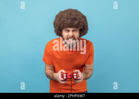 Portrait of crazy funny man with Afro hairstyle grimacing holding joystick, playing video games online, virtual competition. Indoor studio shot isolated on blue background. Stock Photo