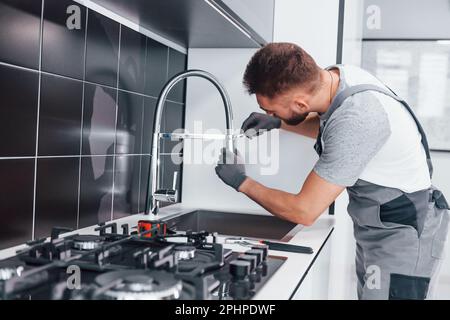 Young professional plumber in grey uniform fixing water tap on the kitchen Stock Photo