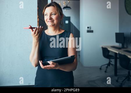 Businesswoman having business call working on tablet in office. Mature busy woman using touch pad computer standing by window in modern interior. Fema Stock Photo