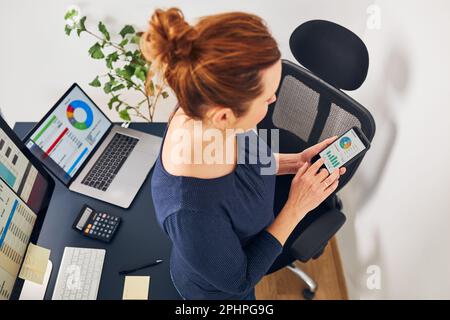 Businesswoman working with financial data on charts and tables on smartphone and computer. Woman entrepreneur standing by desk in office Stock Photo