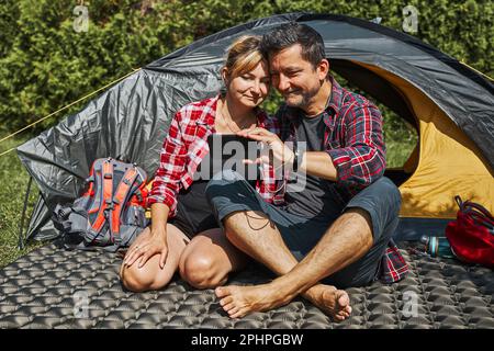 Couple having video call with friends using smartphone while sitting in tent at camping. People relaxing in tent during summer vacation. Actively spen Stock Photo