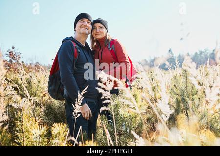 Hugging couple enjoying nature while vacation trip. Hikers with backpacks looking at mountains view. People standing in tall grass on path to mountain Stock Photo