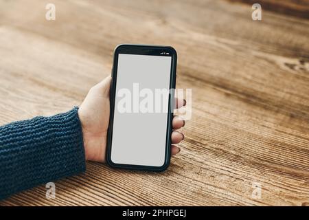 Female hand holding smartphone mockup with solid white color blank empty isolated screen on wooden background. Person using technology. Mobile phone w Stock Photo