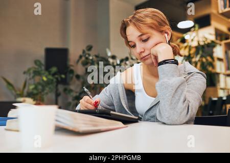 Student learning in university library. Young woman writing essay and making notes using computer tablet. Focused student studying for college exams. Stock Photo