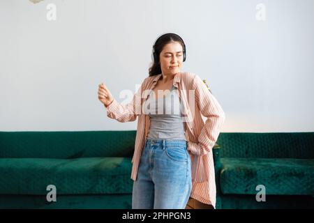 Portrait of young Caucasian pretty woman wearing headphones dancing in living room. Green sofa in background. Home recreation. Stock Photo